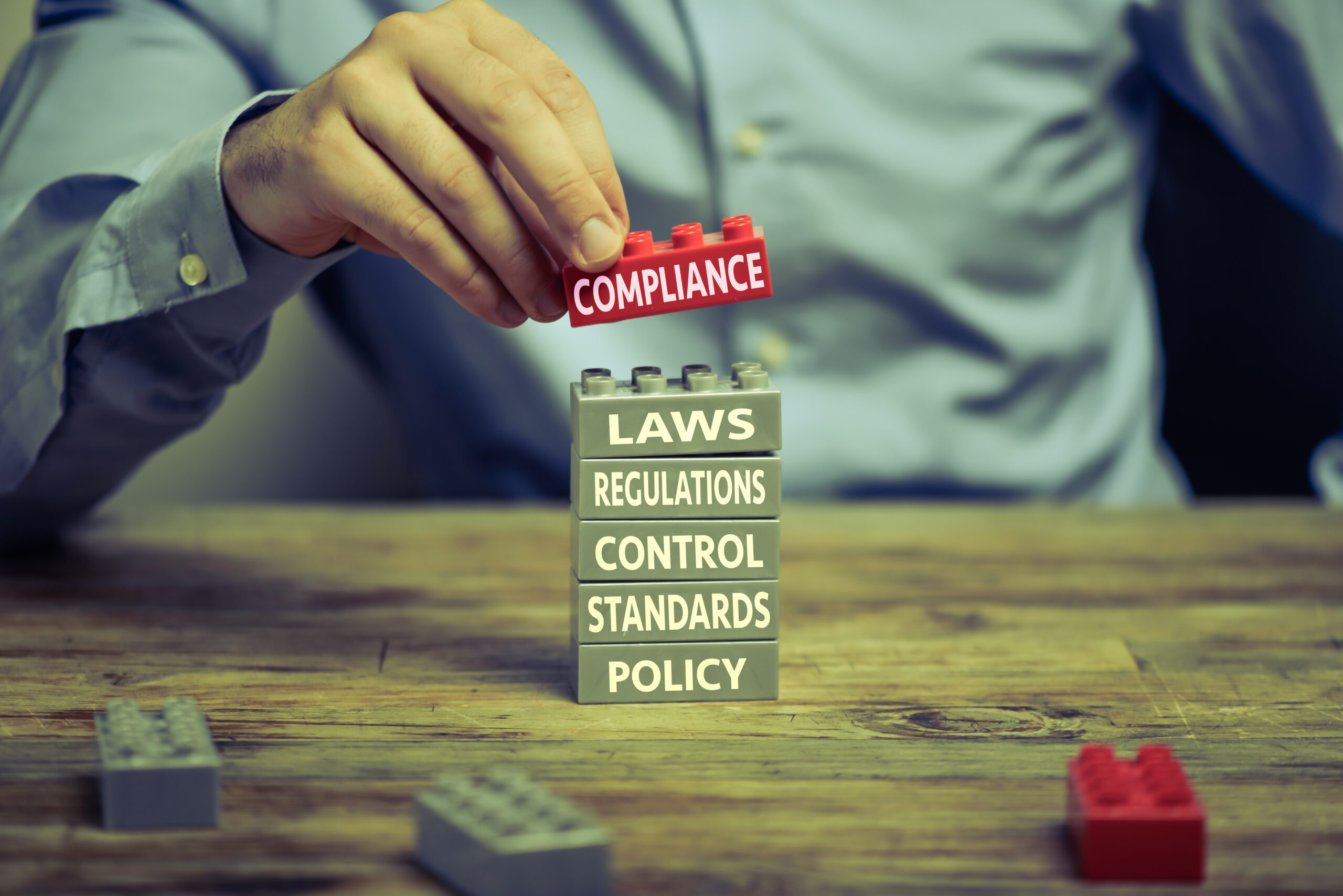 Man in business shirt playing with toy blocks on wood desk. There are compliance related words printed on the blocks