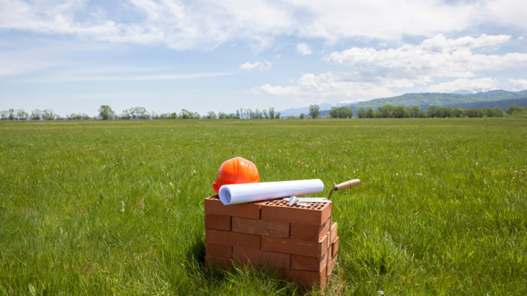 construction hat, building plans sat in top of a pile of bricks in the middle of a field representing green building projects